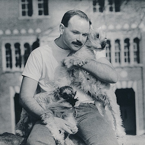 Black and white photograph of person with light skin tone, brown hair, and mustache holding two dogs