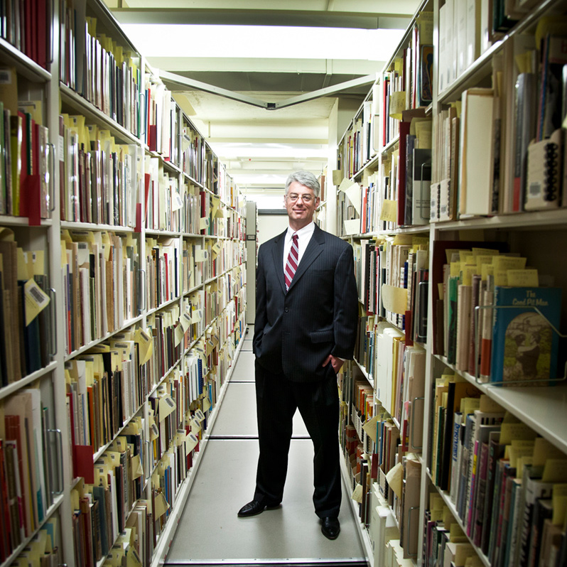 Randy Gue, an adult with light skin tone and white hair, standing between two long library bookshelves