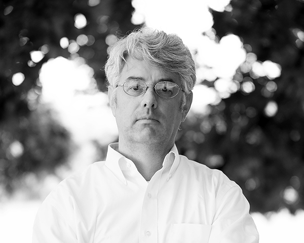 Black and white photograph of curator Randy Gue, an adult with light skin tone, white hair, and wire glasses