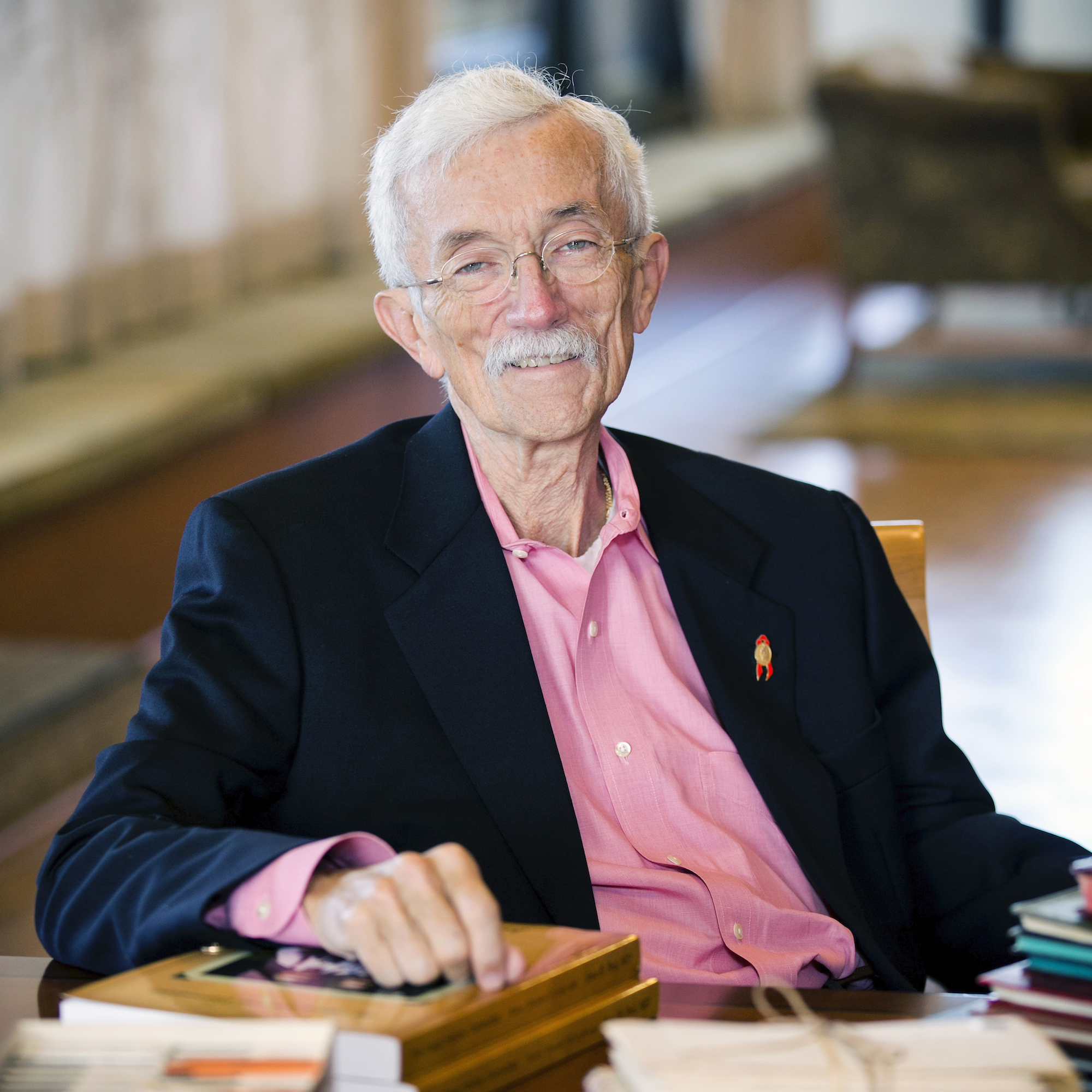 Jesse R. Peel, an older person with light skin tone, white hair and mustache, and wire glasses, seated at book-covered desk