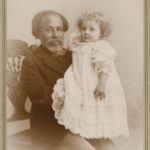 Photograph of older man with dark skin tone holding child with light skin tone