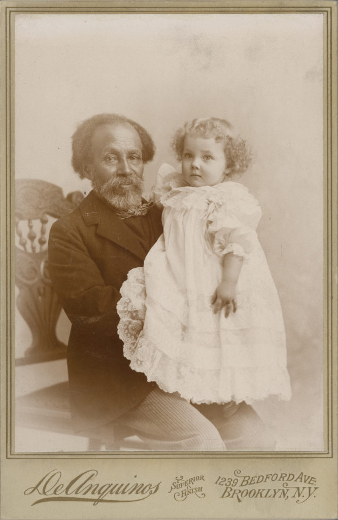 Photograph of older man with dark skin tone holding child with light skin tone